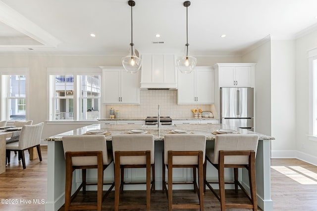 kitchen with decorative backsplash, wood finished floors, stainless steel appliances, premium range hood, and an island with sink