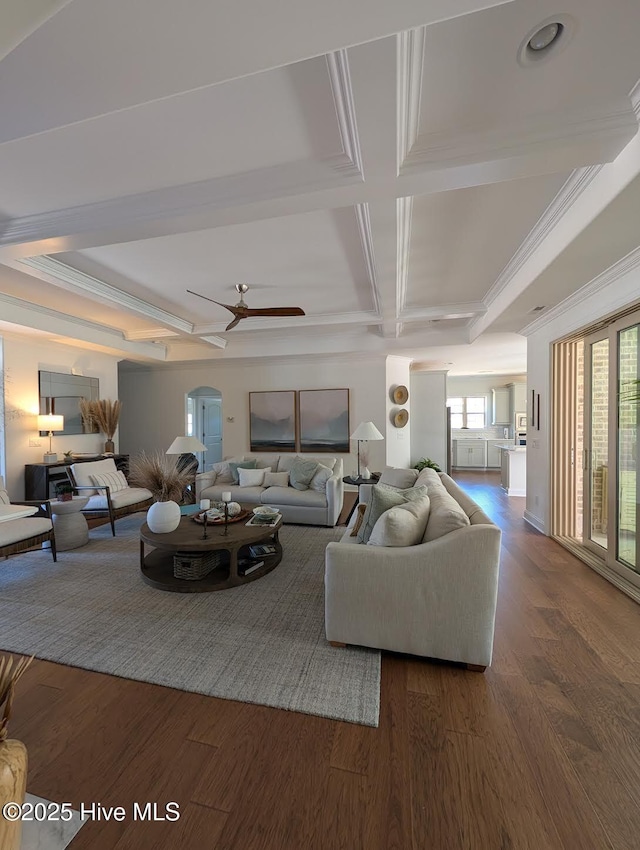 living area with coffered ceiling, a ceiling fan, beamed ceiling, arched walkways, and dark wood-style flooring