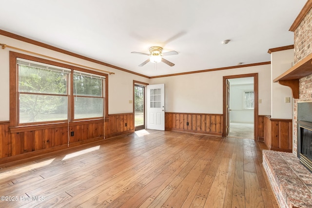 unfurnished living room with crown molding, wainscoting, light wood-style floors, a large fireplace, and a ceiling fan