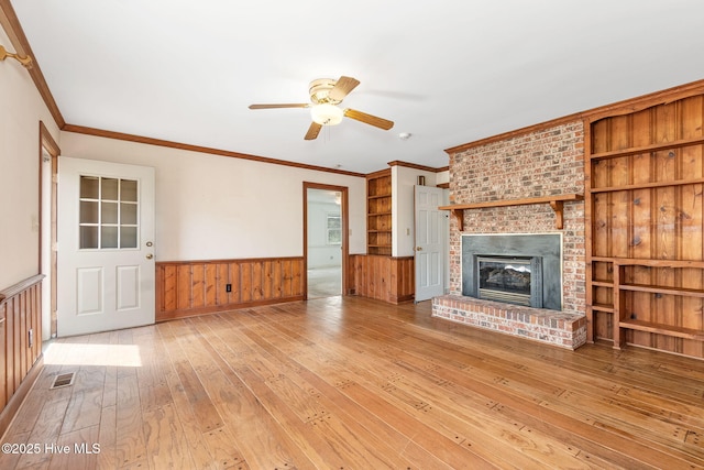 unfurnished living room featuring ceiling fan, crown molding, wainscoting, hardwood / wood-style flooring, and a brick fireplace