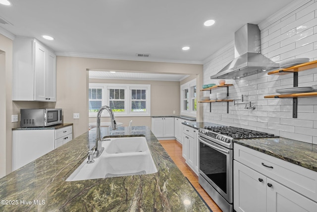 kitchen featuring open shelves, a sink, stainless steel appliances, light wood finished floors, and wall chimney exhaust hood