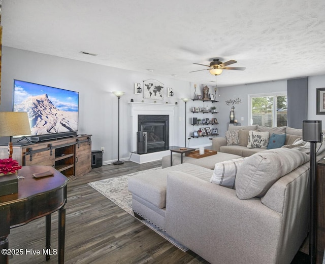 living area featuring a glass covered fireplace, visible vents, a ceiling fan, and dark wood finished floors