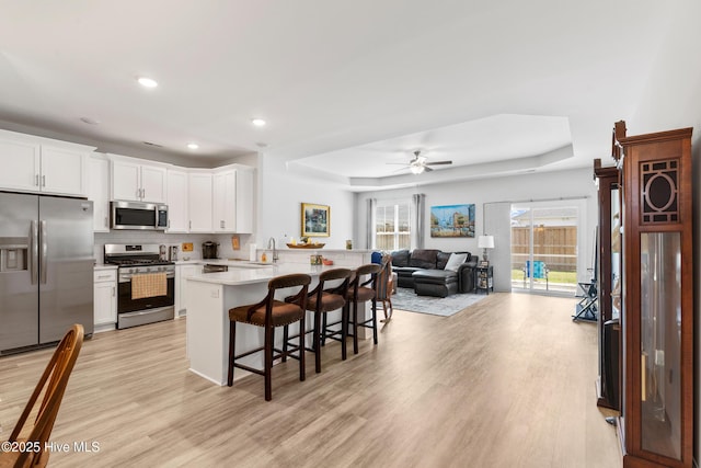 kitchen with a peninsula, light wood-style flooring, a tray ceiling, a kitchen bar, and appliances with stainless steel finishes