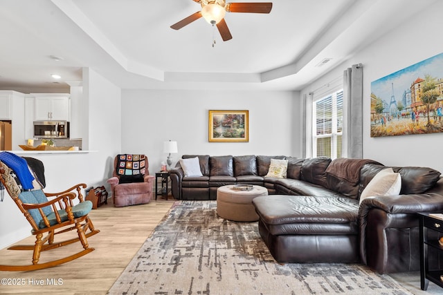 living room featuring a raised ceiling, light wood-type flooring, a ceiling fan, and visible vents