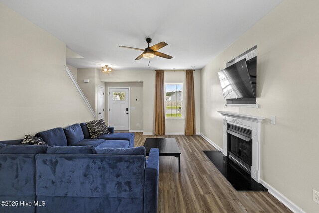 living room featuring dark wood-type flooring, ceiling fan, baseboards, and a glass covered fireplace