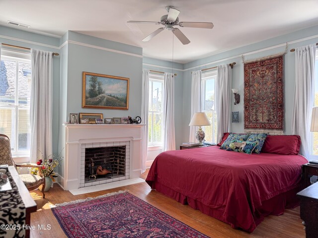 bedroom featuring wood finished floors, a tiled fireplace, visible vents, and a ceiling fan