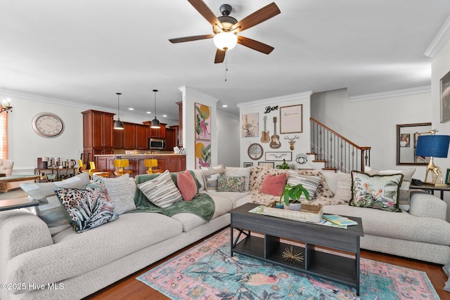 living room with crown molding, ceiling fan with notable chandelier, wood finished floors, and stairway