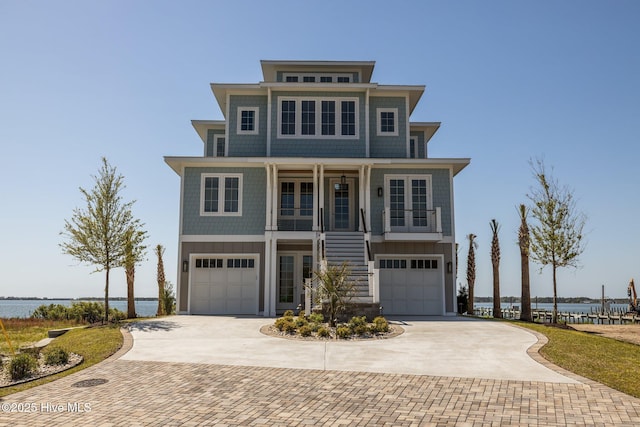 beach home with decorative driveway, a water view, an attached garage, a porch, and stairs