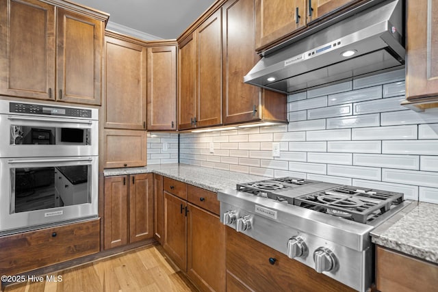 kitchen featuring light wood finished floors, appliances with stainless steel finishes, backsplash, under cabinet range hood, and light stone countertops