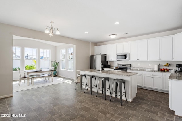 kitchen featuring white cabinets, a notable chandelier, appliances with stainless steel finishes, a center island with sink, and light stone countertops