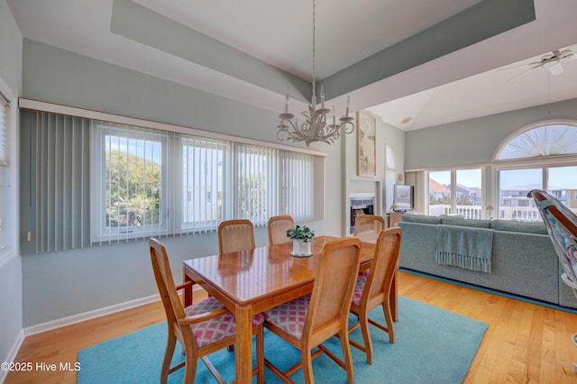 dining room featuring a raised ceiling, light wood-style floors, baseboards, a fireplace, and ceiling fan with notable chandelier