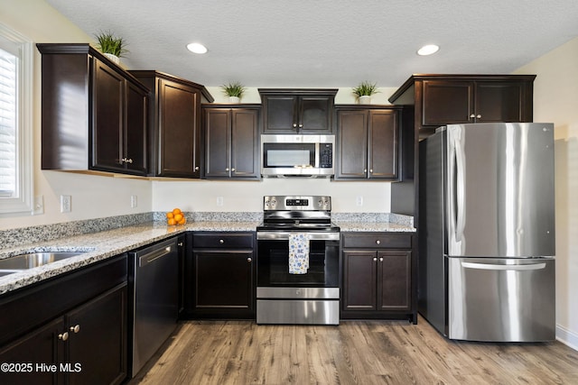 kitchen featuring dark brown cabinetry, light wood-type flooring, light stone counters, and appliances with stainless steel finishes