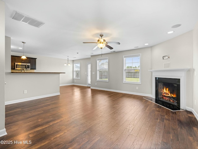 unfurnished living room with ceiling fan, a glass covered fireplace, visible vents, baseboards, and dark wood-style floors