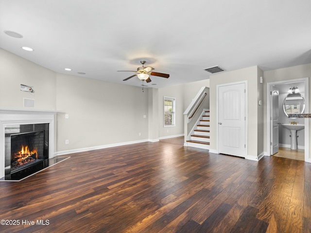 unfurnished living room featuring ceiling fan, a warm lit fireplace, visible vents, stairway, and wood finished floors
