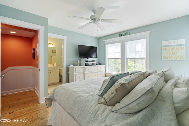 bedroom with ensuite bath, a wainscoted wall, light wood finished floors, and a ceiling fan