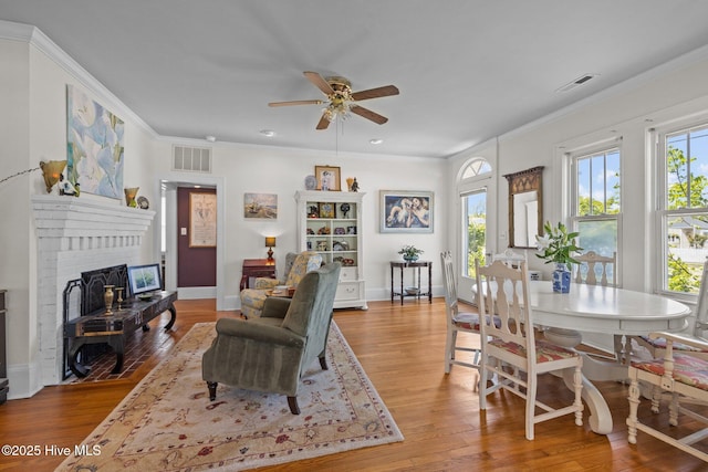 living area with a ceiling fan, visible vents, a brick fireplace, wood finished floors, and ornamental molding