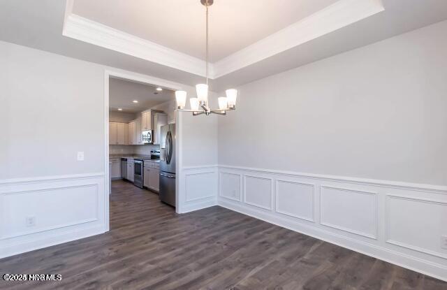 unfurnished dining area with a raised ceiling, a notable chandelier, dark wood finished floors, crown molding, and wainscoting