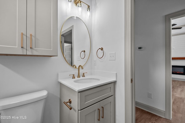 half bathroom with vanity, a glass covered fireplace, wood finished floors, toilet, and baseboards