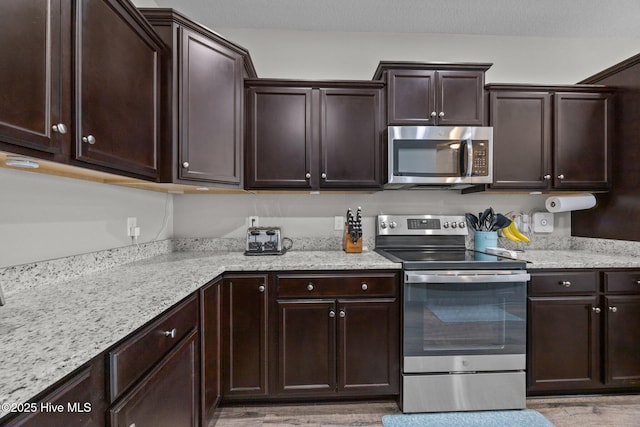 kitchen featuring dark brown cabinets, light stone countertops, appliances with stainless steel finishes, and light wood finished floors