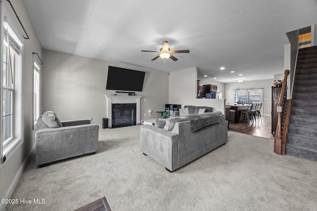 carpeted living area featuring baseboards, a ceiling fan, a glass covered fireplace, and stairway