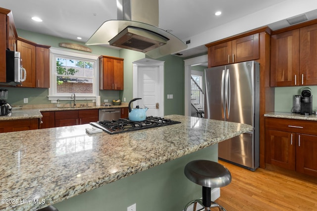 kitchen with a sink, appliances with stainless steel finishes, a kitchen bar, light stone countertops, and island range hood