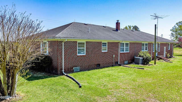 rear view of property featuring a yard, roof with shingles, brick siding, a chimney, and central AC unit