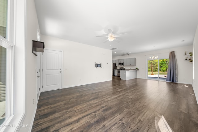 unfurnished living room with ceiling fan with notable chandelier, baseboards, and dark wood-style floors