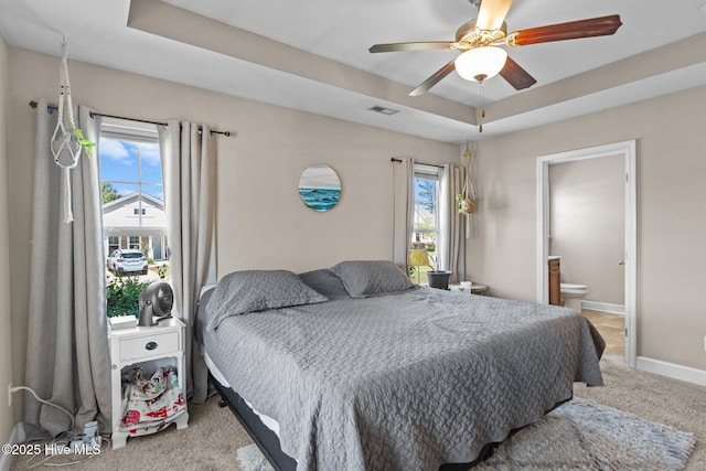 carpeted bedroom with a tray ceiling, ensuite bathroom, visible vents, and multiple windows