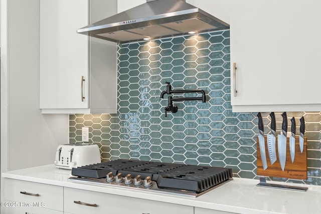 kitchen with wall chimney exhaust hood, stainless steel gas stovetop, white cabinetry, light countertops, and backsplash