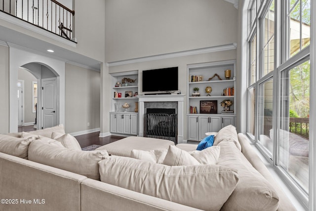 living area featuring a fireplace, arched walkways, a towering ceiling, baseboards, and built in shelves