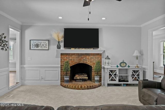 carpeted living room with crown molding, ceiling fan, a fireplace, and wainscoting