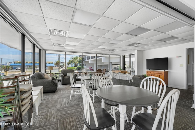 carpeted dining space with plenty of natural light, a drop ceiling, a wall of windows, and baseboards