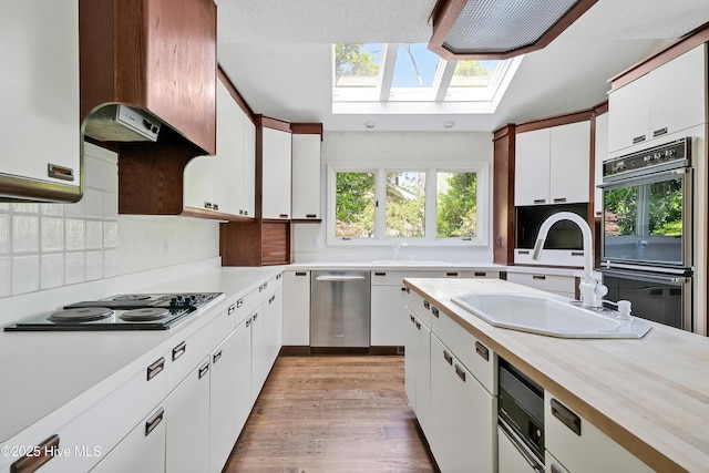 kitchen featuring stainless steel dishwasher, black electric stovetop, a sink, multiple ovens, and a skylight