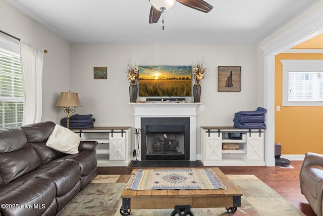 living area featuring a fireplace with raised hearth, baseboards, a ceiling fan, and wood finished floors
