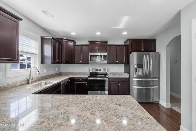 kitchen with dark brown cabinetry, dark wood-style flooring, arched walkways, a sink, and stainless steel appliances