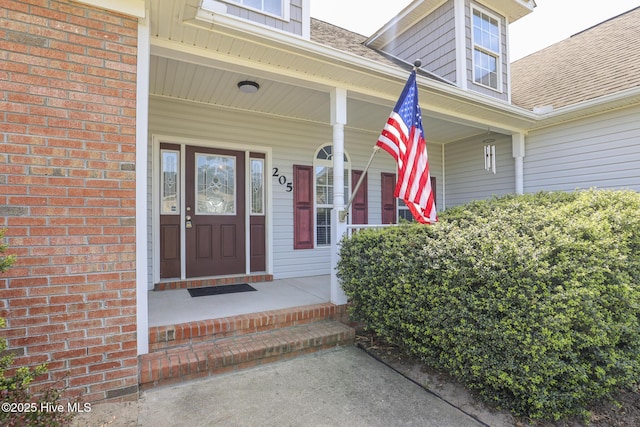 view of exterior entry with a shingled roof, covered porch, and brick siding
