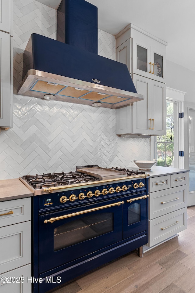 2592 St James Dr SE, Southport, NC 28461 - kitchen featuring range with two ovens, wall chimney exhaust hood, light wood-type flooring, tasteful backsplash, and glass insert cabinets