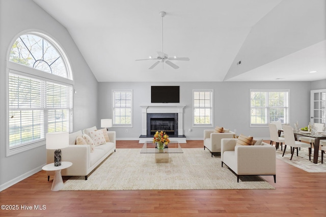 living room with high vaulted ceiling, a ceiling fan, a glass covered fireplace, wood finished floors, and baseboards