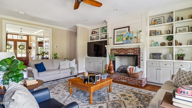 living area featuring wood finished floors, built in shelves, ceiling fan, ornamental molding, and a brick fireplace