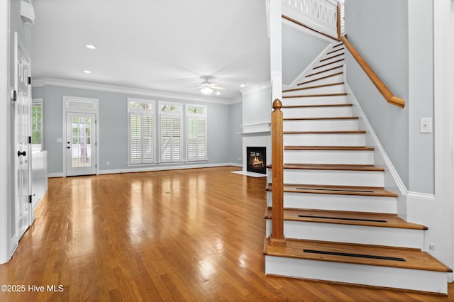 stairway featuring a fireplace with flush hearth, crown molding, ceiling fan, baseboards, and wood finished floors