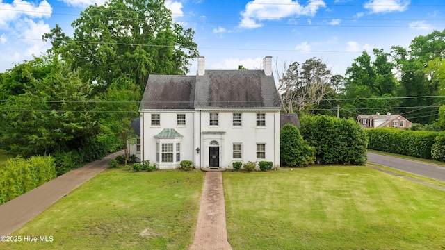 colonial inspired home featuring stucco siding, roof with shingles, a chimney, and a front yard