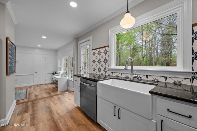 kitchen featuring stainless steel dishwasher, crown molding, dark stone countertops, light wood finished floors, and hanging light fixtures