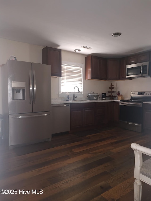 kitchen featuring stainless steel appliances, a sink, dark brown cabinetry, light countertops, and dark wood-type flooring