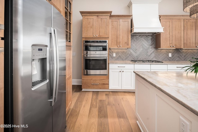 kitchen featuring custom exhaust hood, tasteful backsplash, glass insert cabinets, light wood finished floors, and appliances with stainless steel finishes