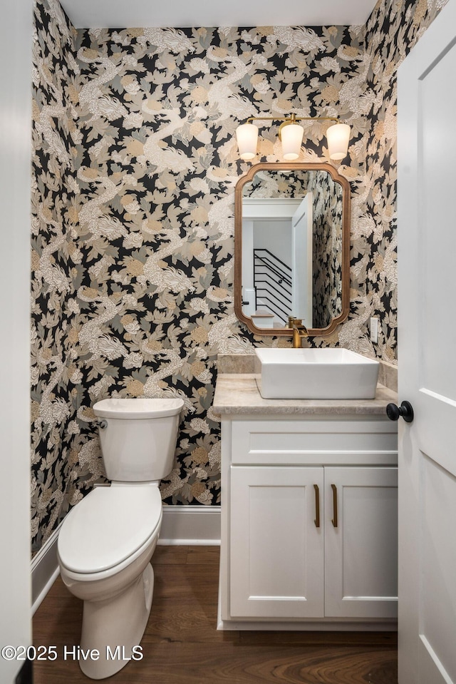 half bath with toilet, baseboards, wallpapered walls, wood finished floors, and vanity