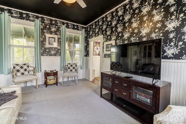 living area featuring wainscoting, wallpapered walls, ceiling fan, ornamental molding, and carpet floors