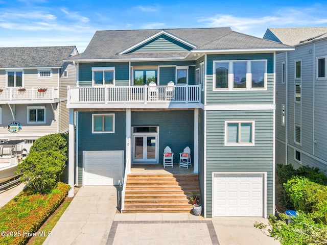 coastal inspired home featuring a shingled roof, an attached garage, a balcony, french doors, and driveway