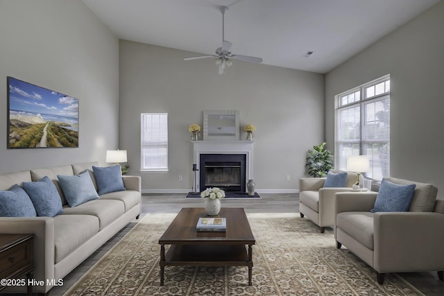 living room featuring wood finished floors, high vaulted ceiling, a ceiling fan, a glass covered fireplace, and baseboards