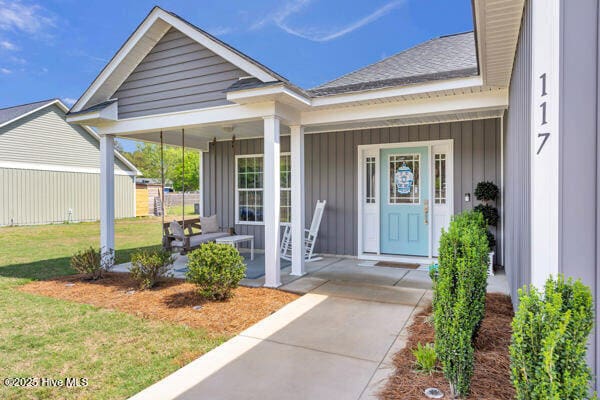 doorway to property featuring a lawn, covered porch, board and batten siding, and a shingled roof