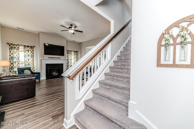stairway featuring baseboards, a ceiling fan, wood finished floors, and a fireplace